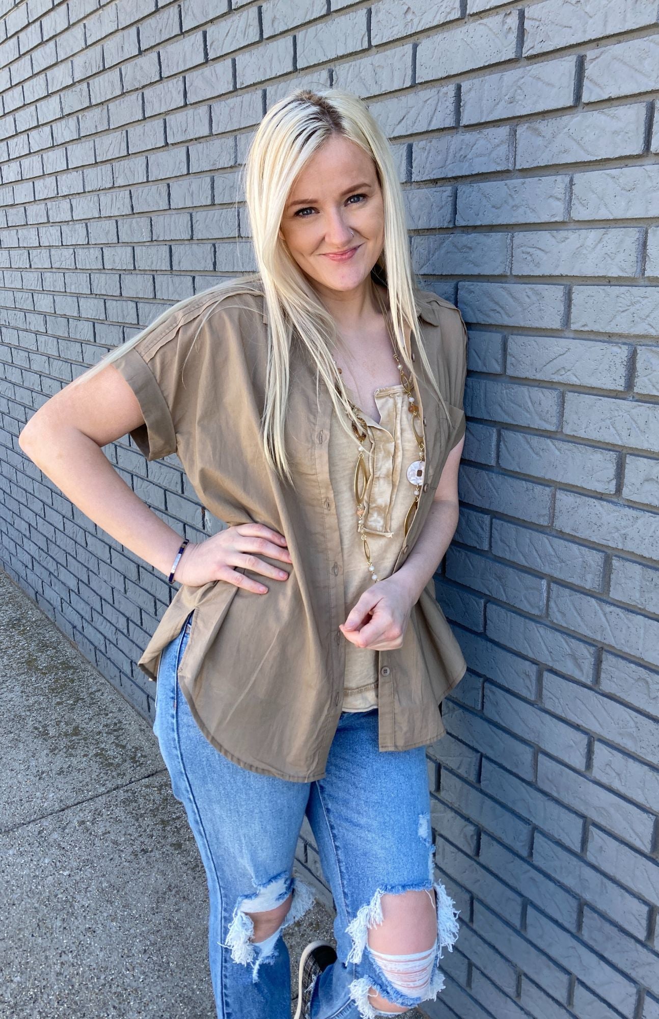 Fashion photo of a young girl with blonde hair. She is wearing a mustard colored tank top with aa brown short sleeve unbuttoned shirt (like a shacket). She is also wearing a pair of light wash distressed jeans.