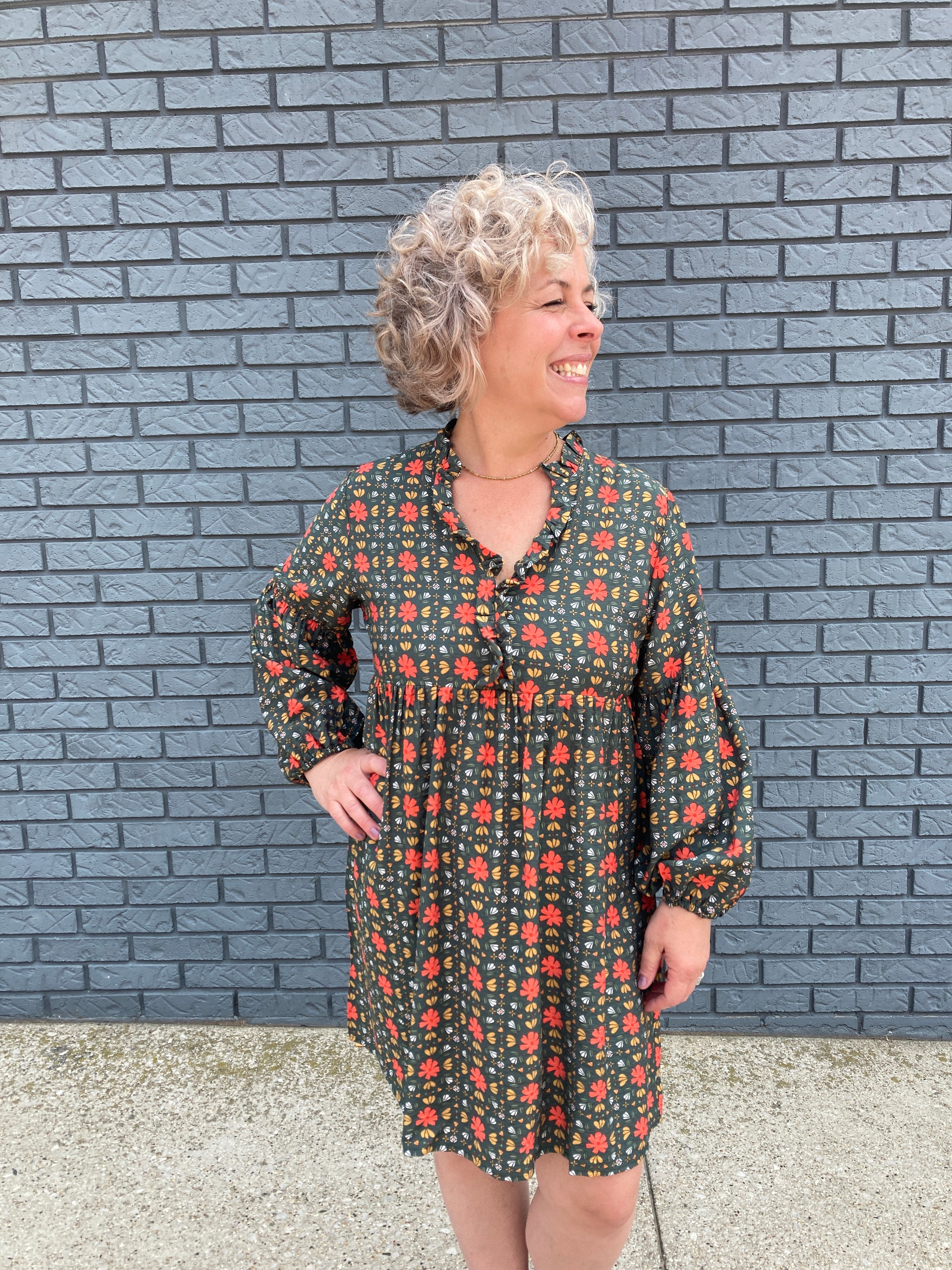 A lovely 50 something woman wearing an army green dress with a vintage floral pattern, puffy sleeves and a ruffled neckline. 