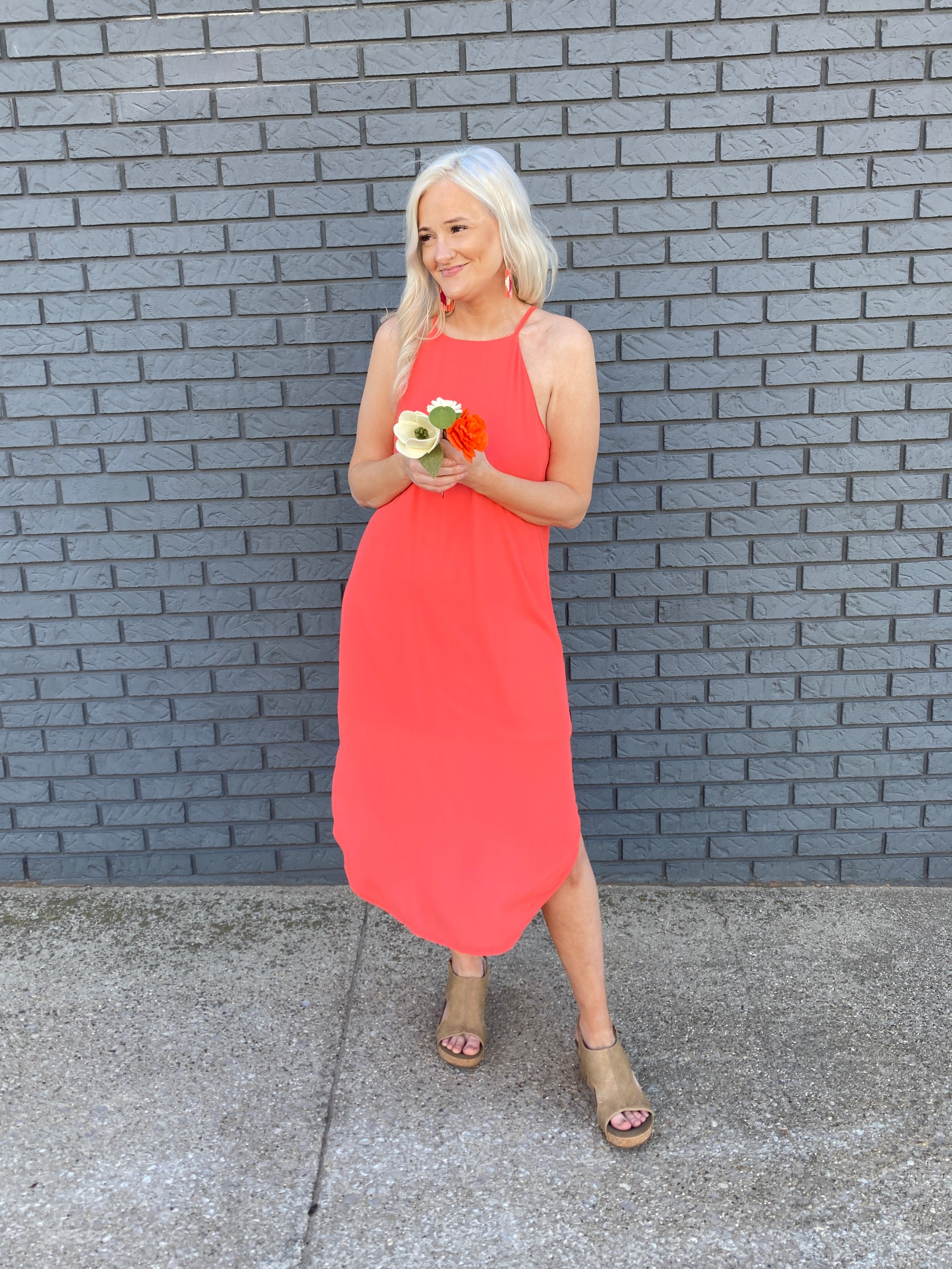  A young woman standing gracefully in a vibrant orange dress. The dress features spaghetti straps, a high neckline, and a maxi length, reaching down to her ankles. The dress has a stylish rounded hem, creating a flowing and elegant silhouette.