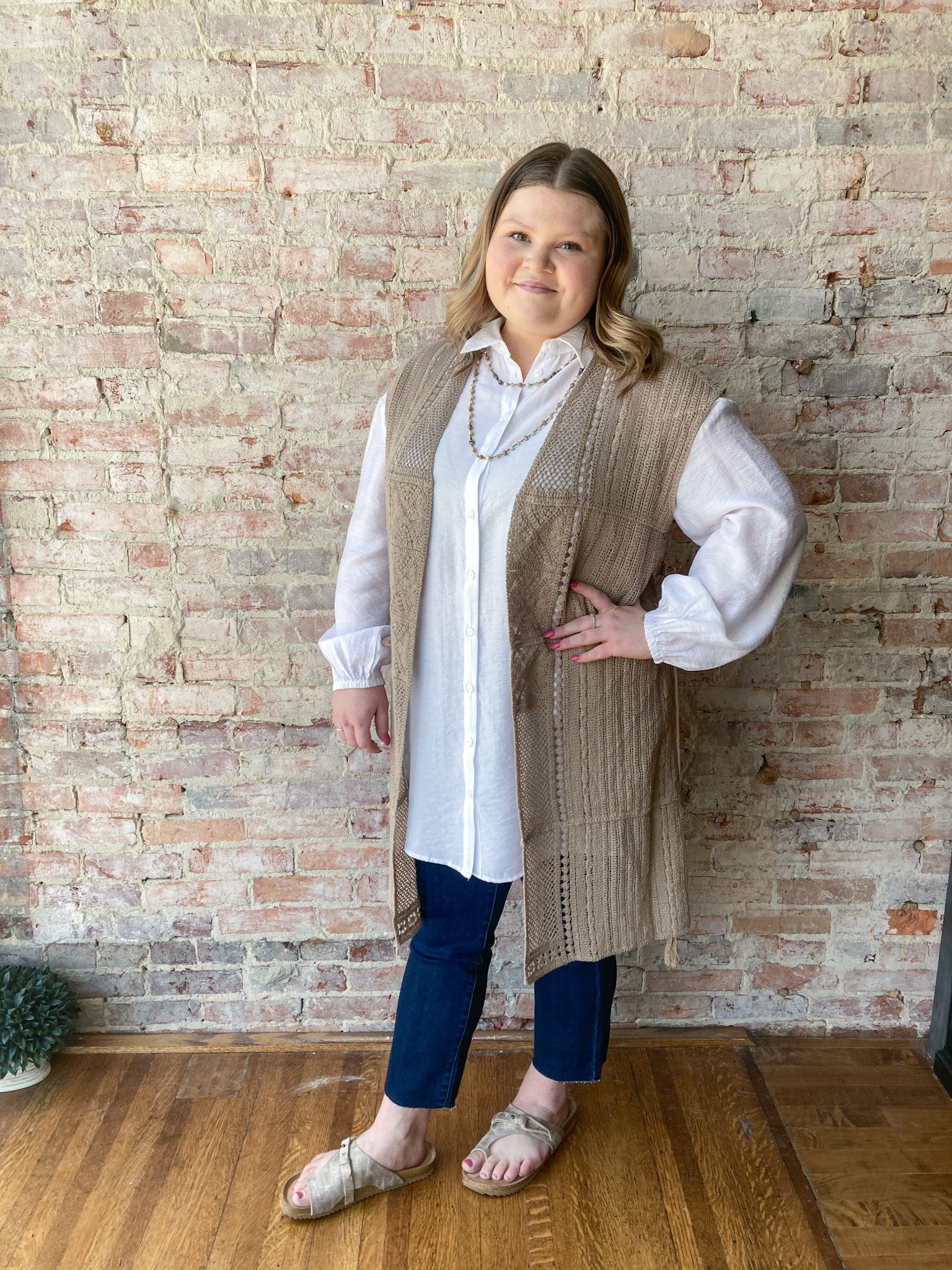 A gorgeous plus size woman wearing a classic oversized extra long white blouse with a tan crochet vest and dark wash jeans. She is also wearing a necklace and some fun sandals.