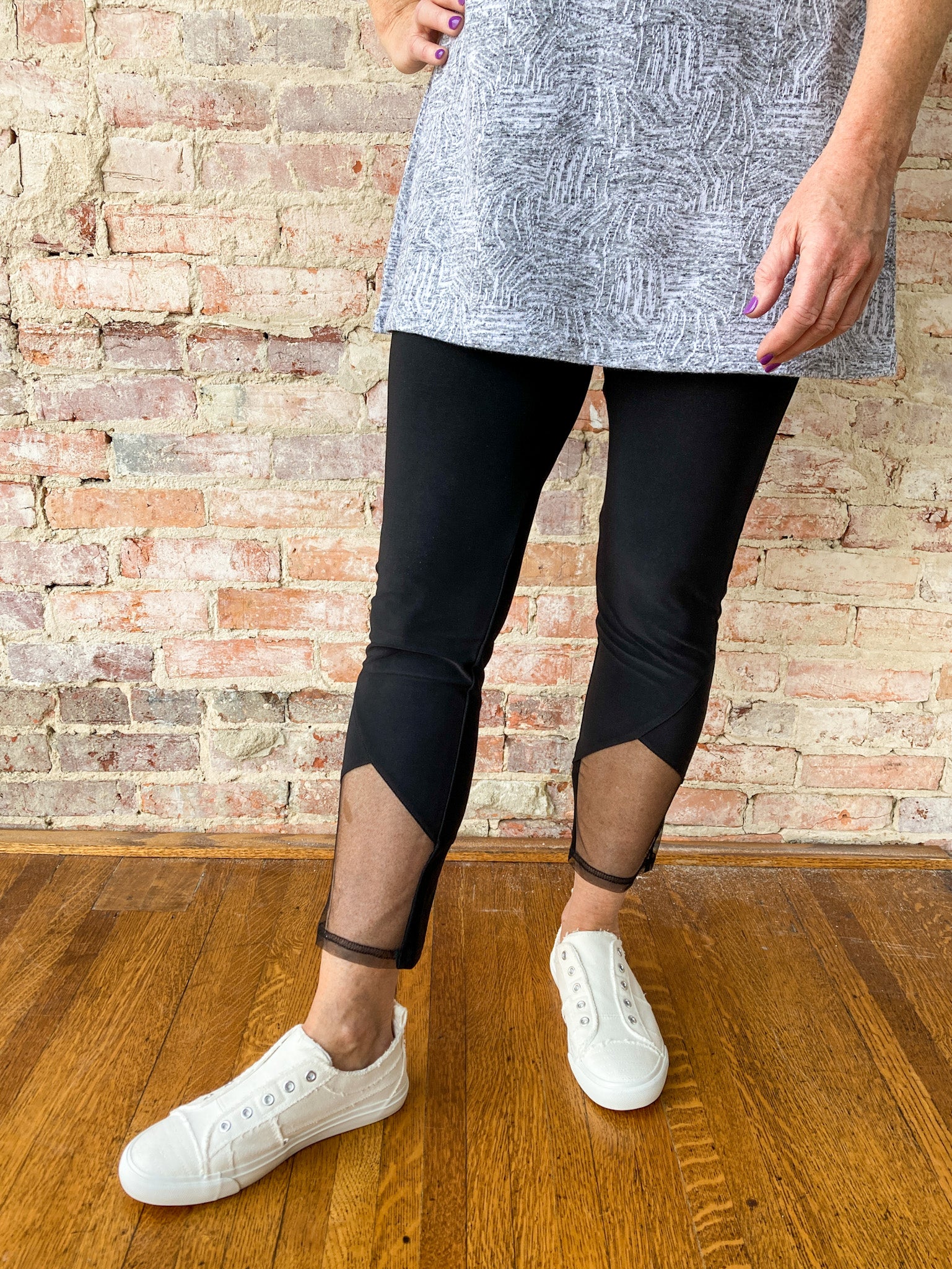 A close up of a woman wearing black leggings with a peek-a-boo mesh design in the front. She is also wearing a grey tunic and some white babalu shoes