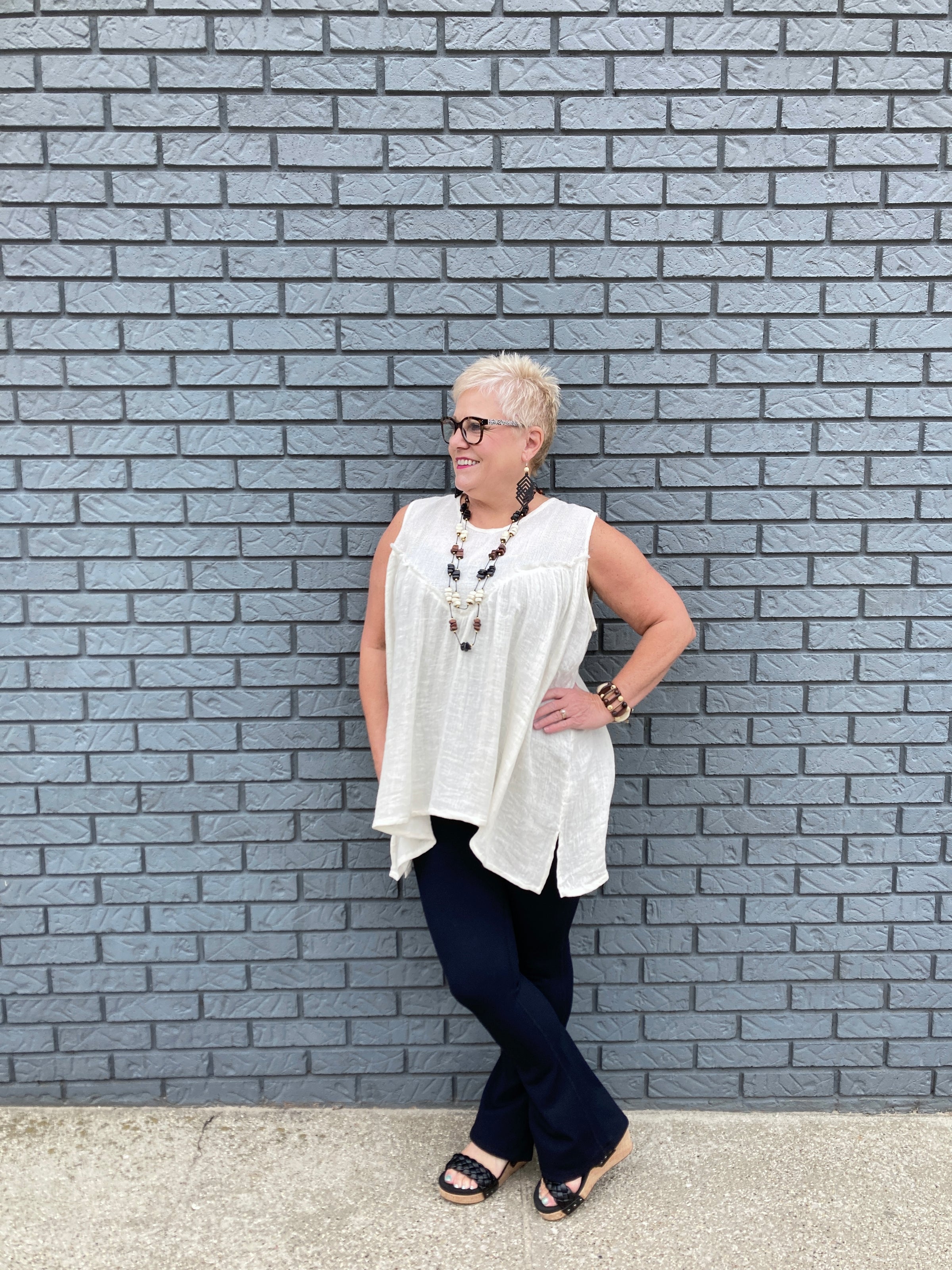 Fashion photo of a woman in her 50s with a pixie haircut. She has bleach blonde hair and is wearing a cream colored babydoll tank top with black flared leggings and black sandals. She looks very chic and trendy.