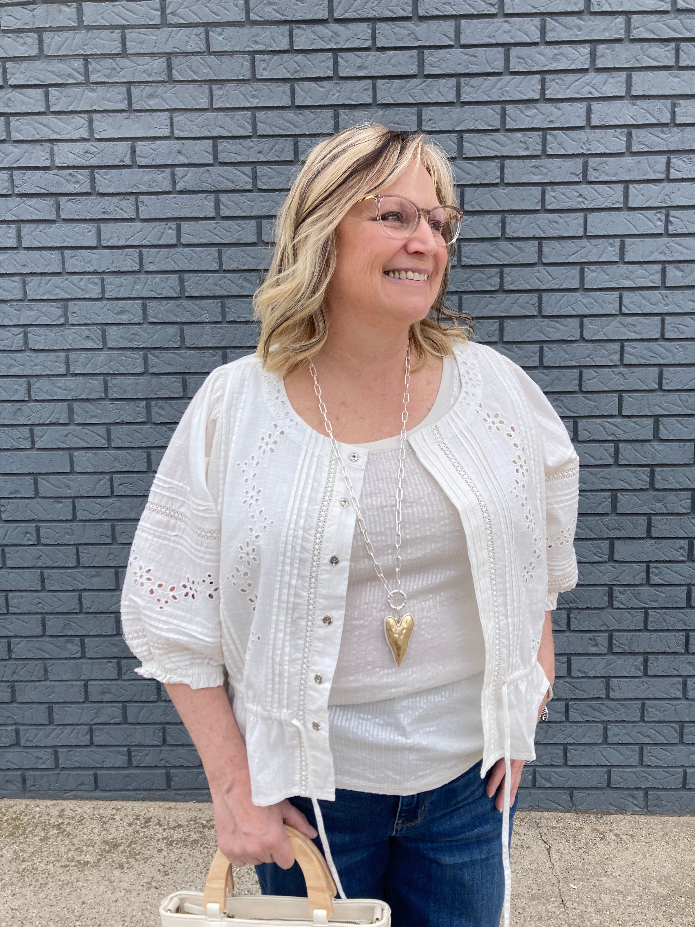 A blonde professional woman in her 50's wearing a chic white peasant blouse as a shacket over a white tank top. She is also wearing blue jeans and a gold heart necklace in Cadillac Michigan