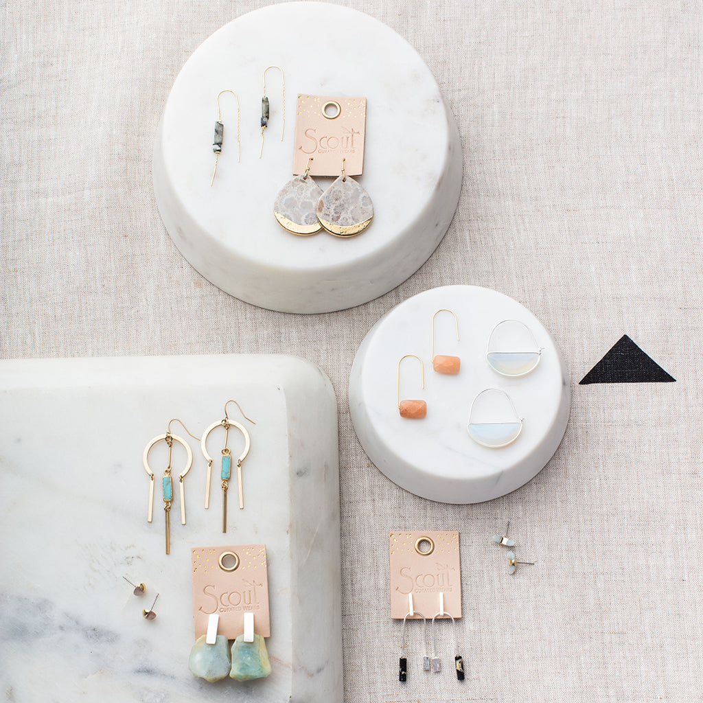 Scout brand earrings collection displayed on white marble platforms showing various stone and gold designs