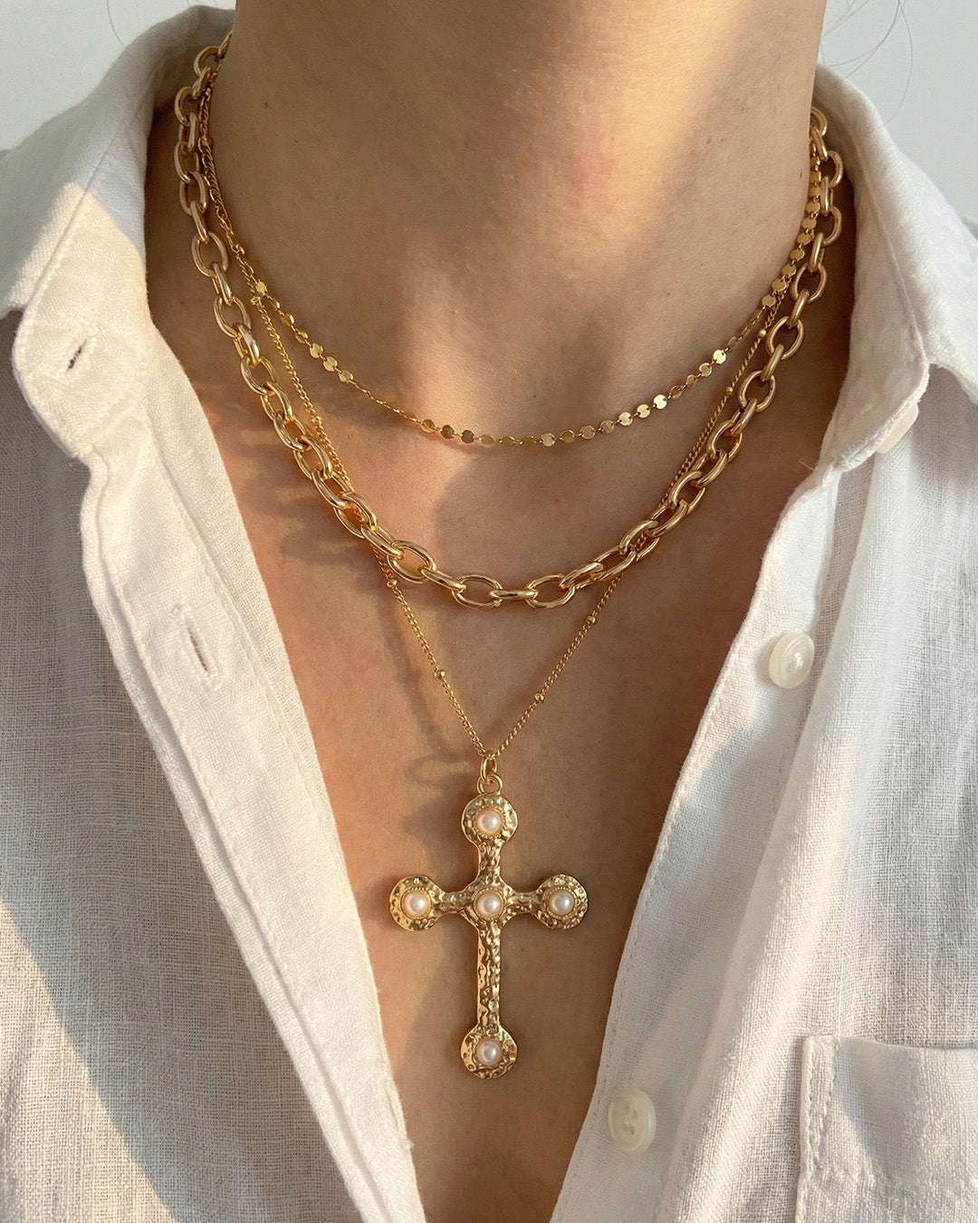 Three layered gold necklaces featuring a hammered cross pendant with pearl embellishments worn on neck
