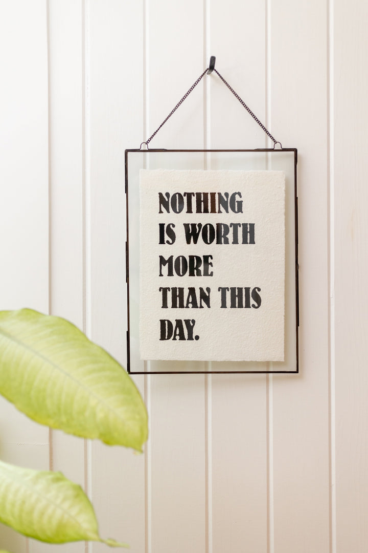 Framed handmade paper print reading 'Nothing Is Worth More Than This Day' hung on white wall