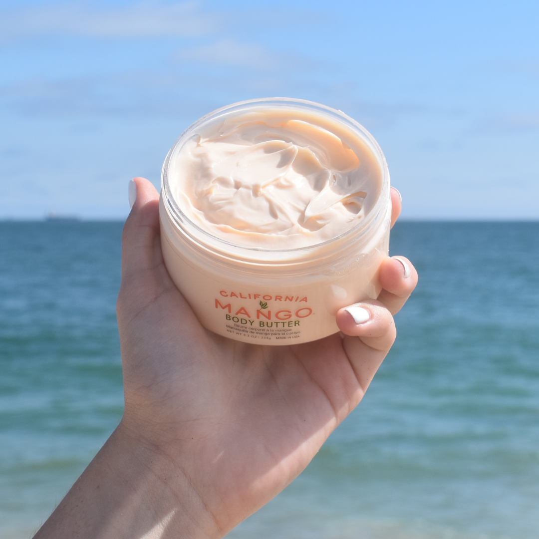 California Mango Body Butter 8.3oz jar held open against a beach background, showing creamy peach product.