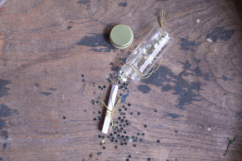 Clear glass wishing bottle ornament with gold star confetti scattered on wooden surface