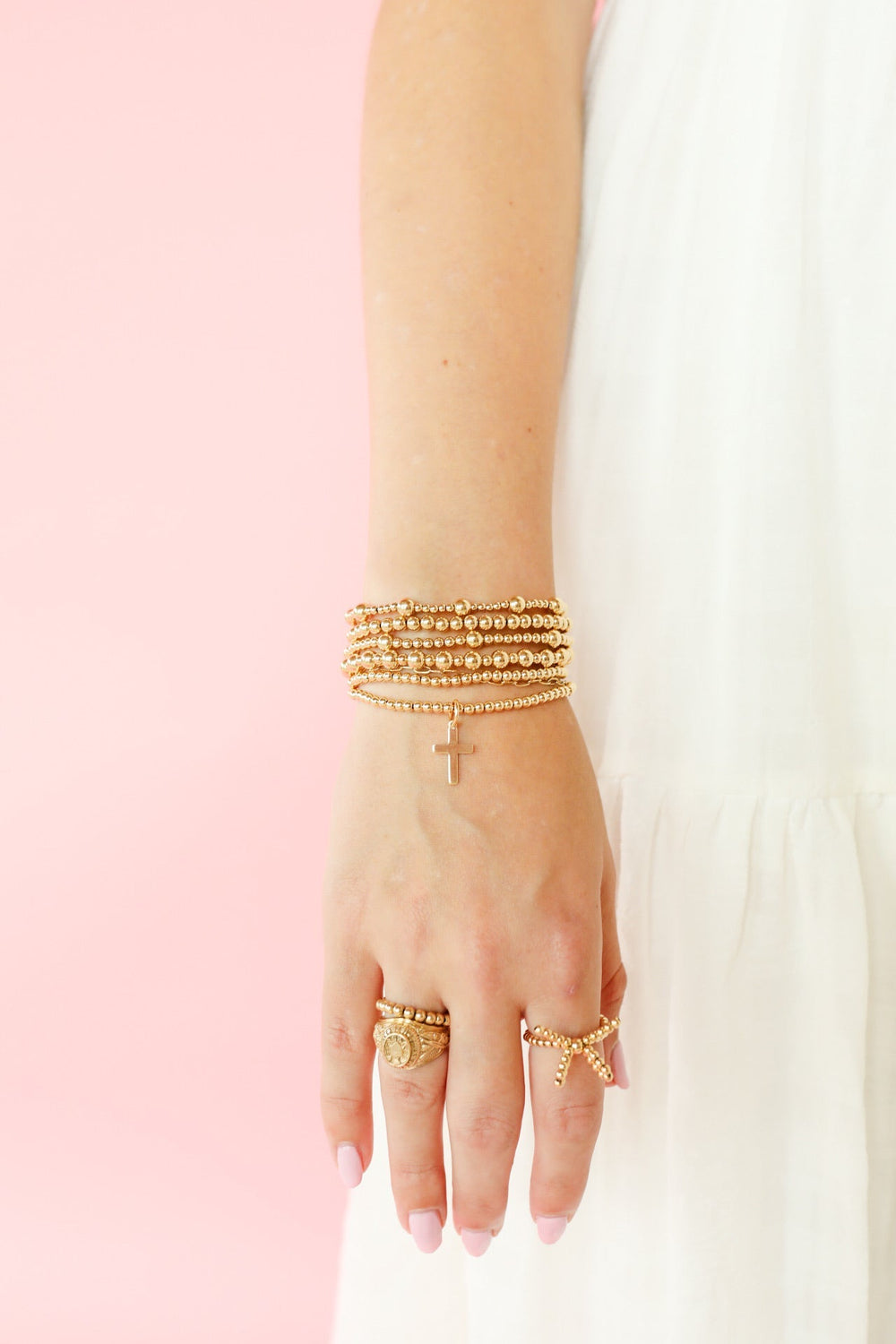 Gold beaded bracelet stack with mini cross charm worn on wrist with gold rings