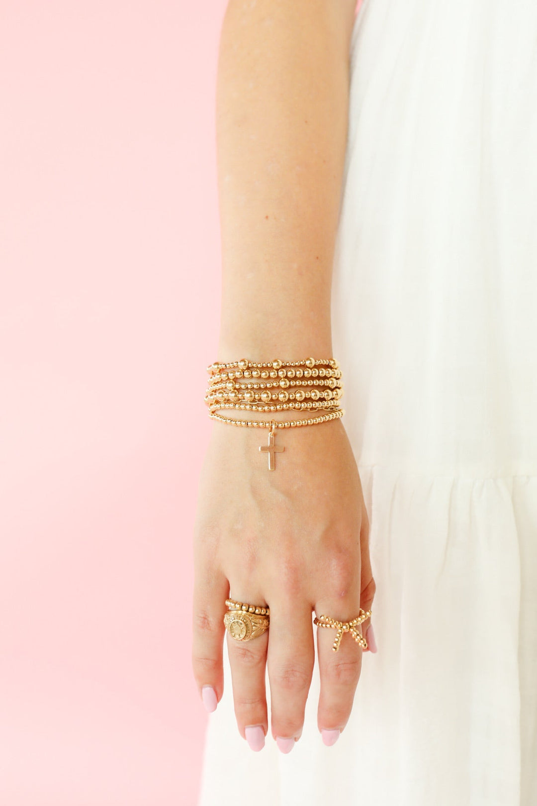 Gold beaded bracelet stack with mini cross charm worn on wrist with gold rings