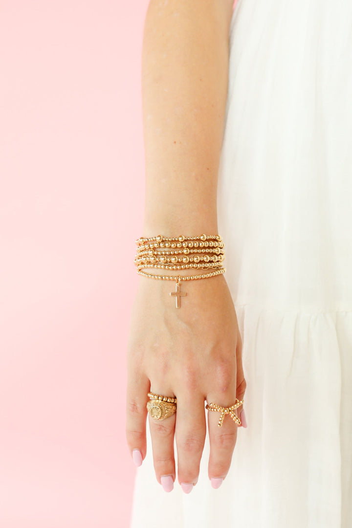 Gold beaded bracelet stack with mini cross charm worn on wrist with gold rings