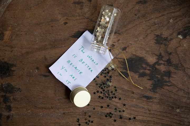 Glass wishing bottle ornament with gold star confetti and handwritten inspirational note on wood surface