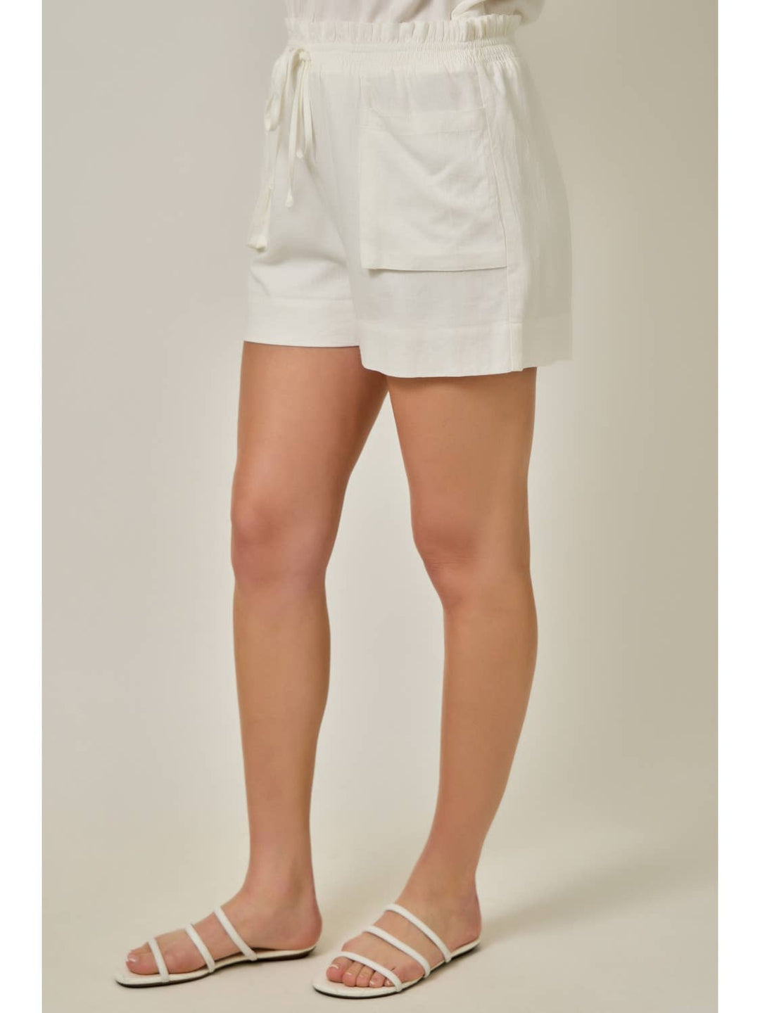 Model wearing ivory linen shorts with elastic waistband and front pockets, paired with white sandals