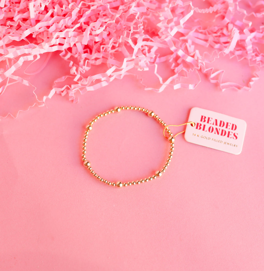 Beaded Blondes Olive Bracelet in 14K gold-filled beads on pink background with tag.