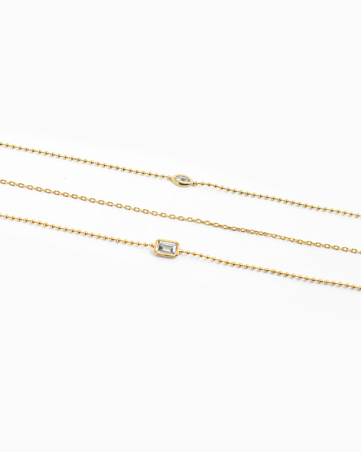 Three layered gold necklaces featuring bezel-set marquise and baguette CZ pendants on delicate chains