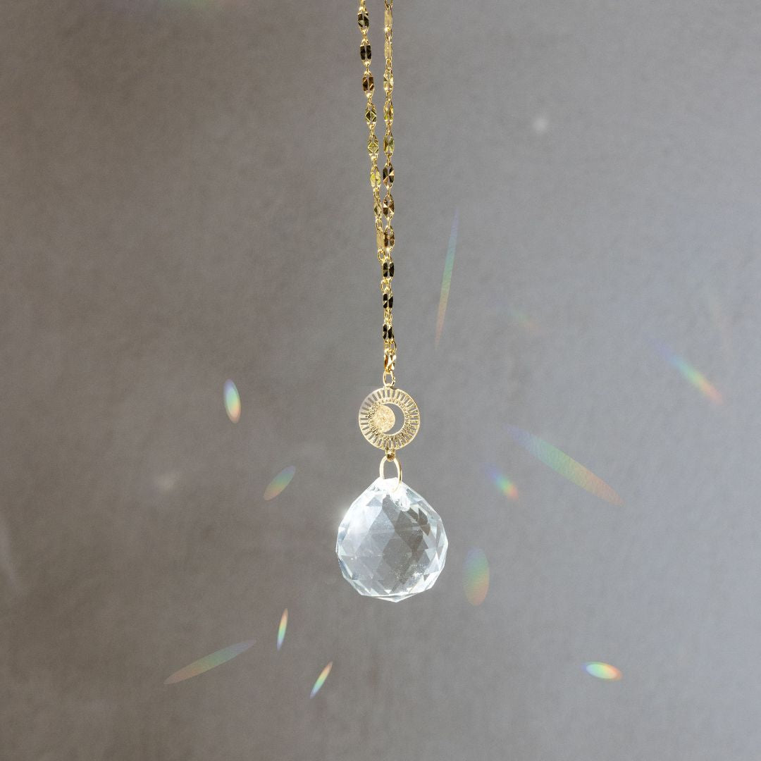 Scout Pocket Suncatcher with clear crystal prism, gold chain, and radiating moon medallion creating rainbow light reflections