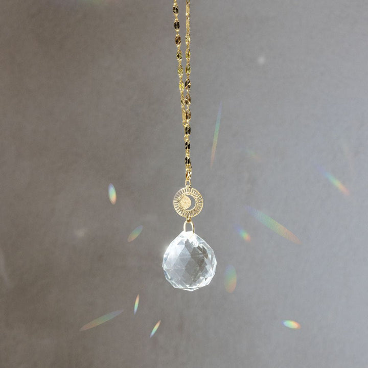 Scout Pocket Suncatcher with clear crystal prism, gold chain, and radiating moon medallion creating rainbow light reflections