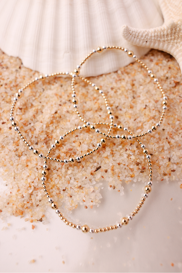 Dainty beaded bracelets with mixed gold and silver beads arranged on sand near seashell