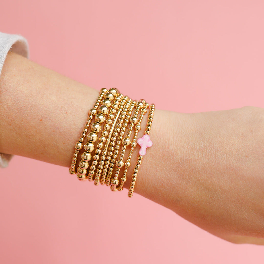 Stacked gold beaded bracelets on wrist featuring Olive Bracelet with pink cross charm