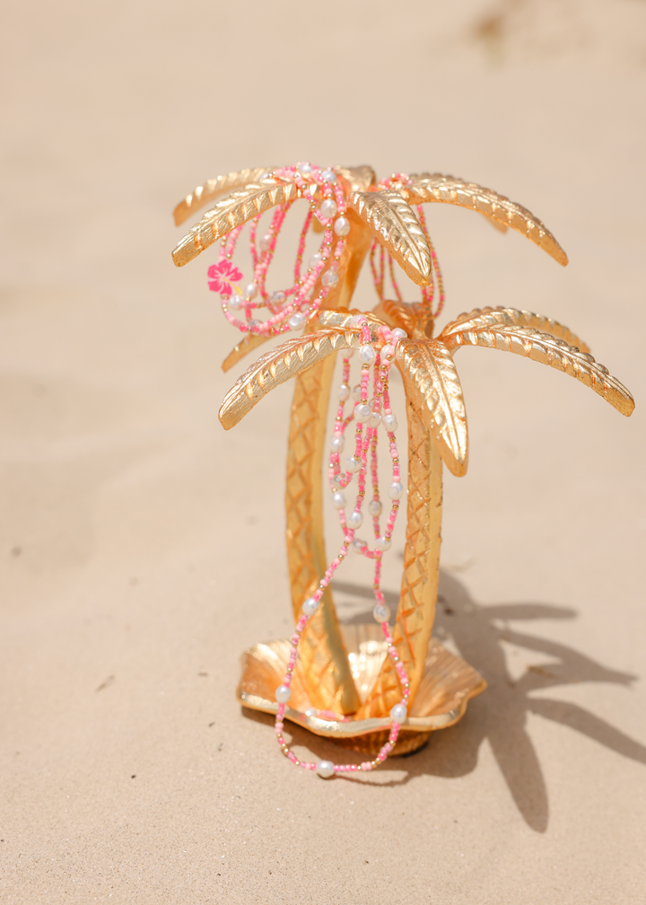 Pink and white beaded Flamingo Sprinkle Bracelet draped on gold palm tree jewelry stand on sand