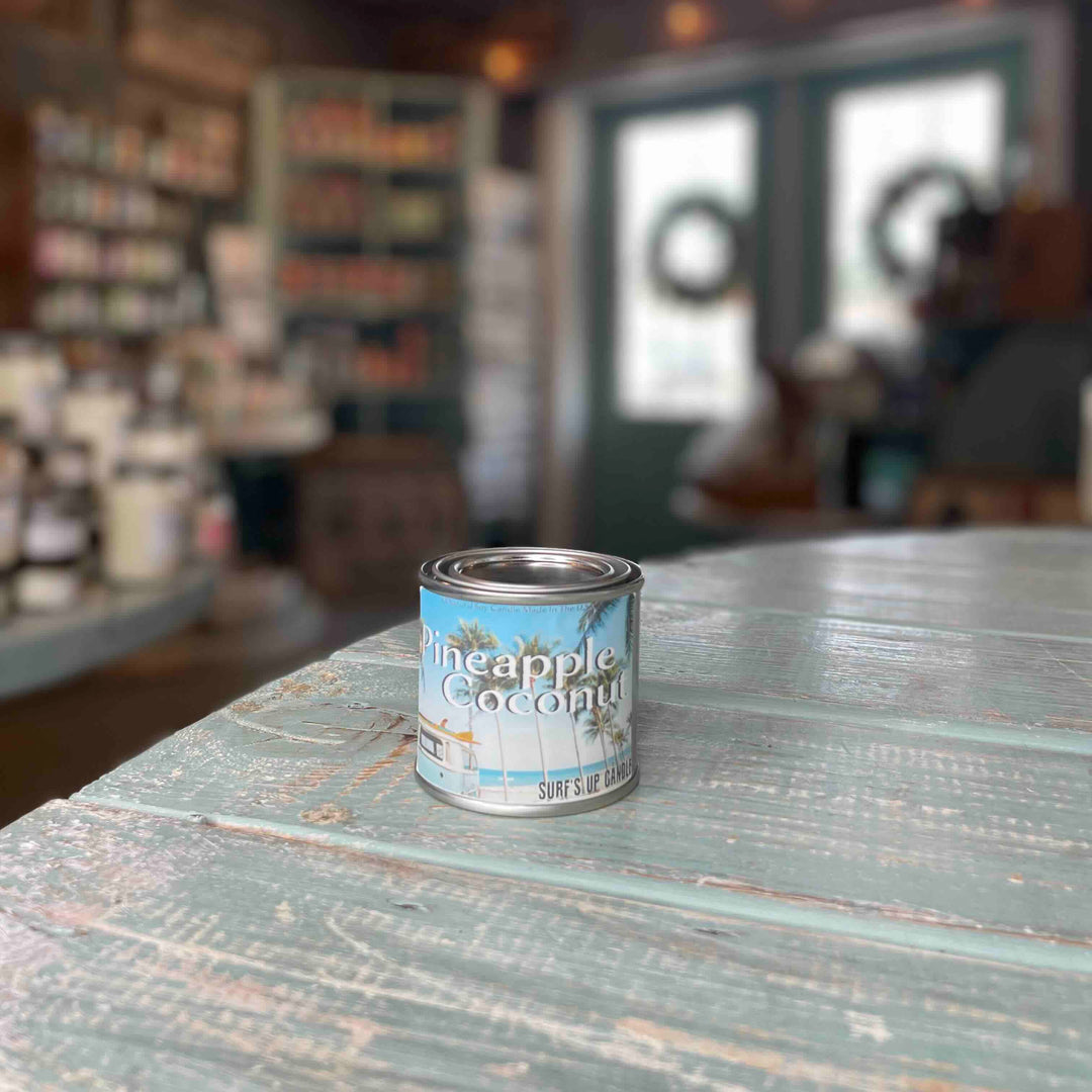 Surfs Up Candle Pineapple Coconut scented paint can candle sitting on a rustic teal wooden surface in a shop.