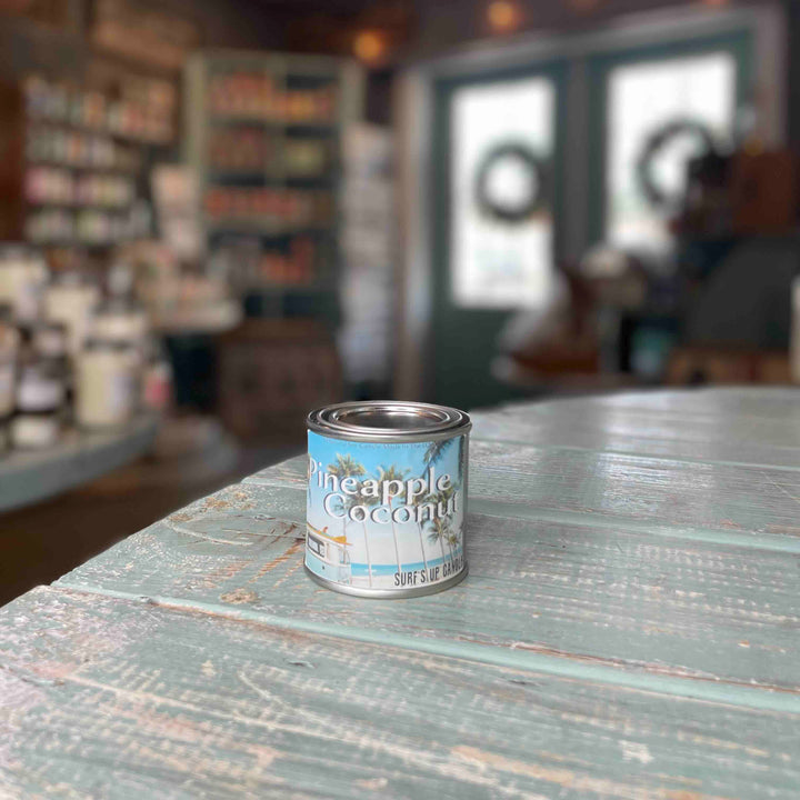 Surfs Up Candle Pineapple Coconut scented paint can candle sitting on a rustic teal wooden surface in a shop.