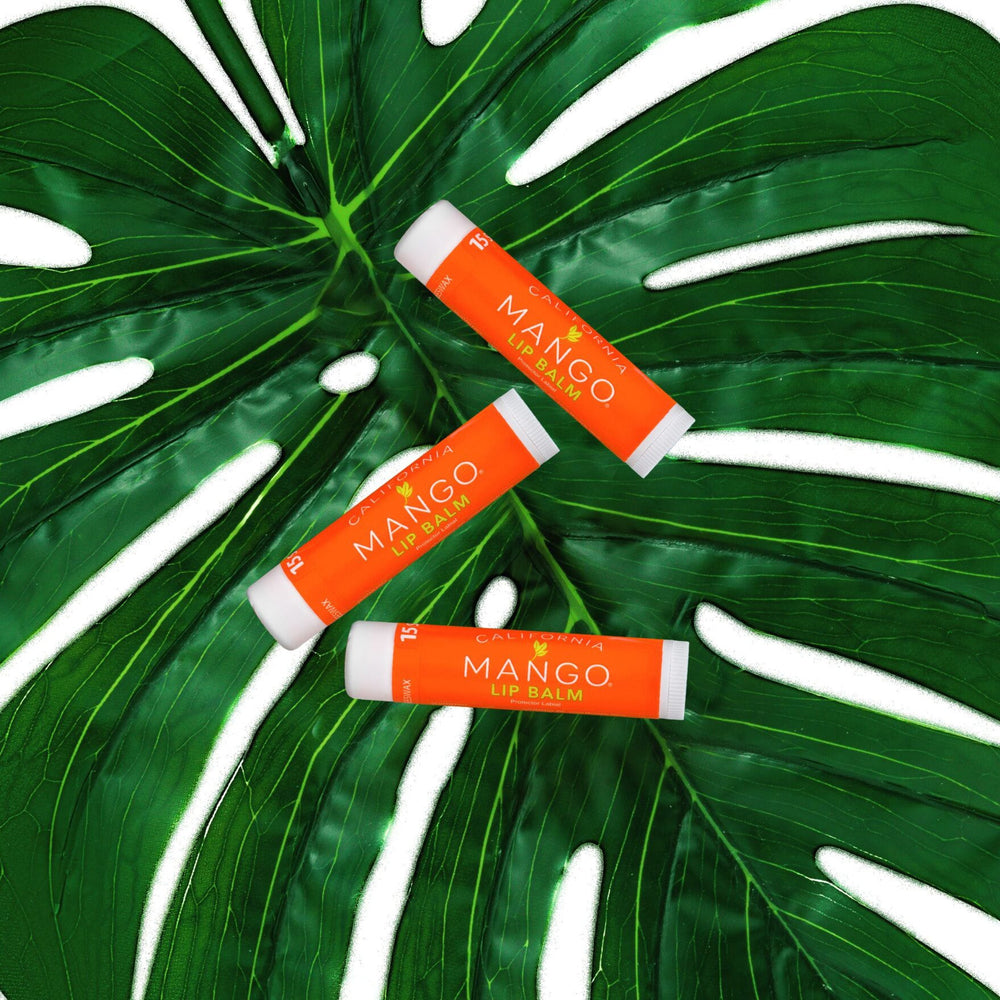 Three California Mango Lip Balm sticks with orange labels arranged on a tropical monstera leaf