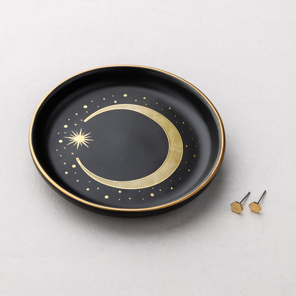 Black ceramic jewelry dish with gold crescent moon design and two small gold hexagon stud earrings