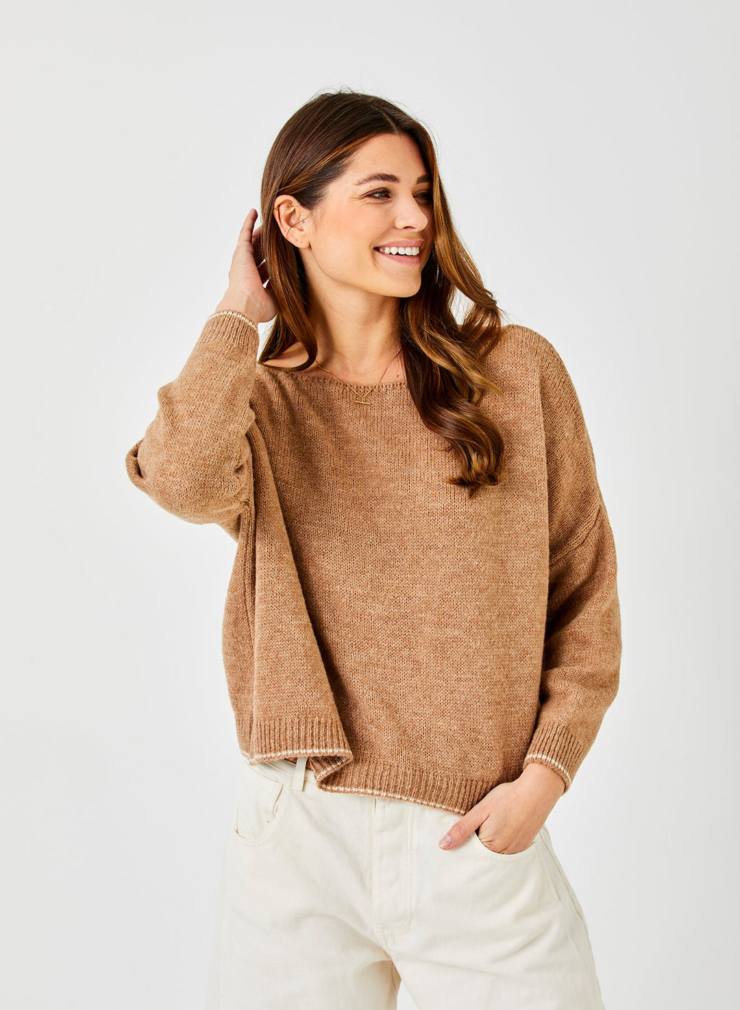 Woman wearing camel colored oversized knit sweater with white pants on neutral background