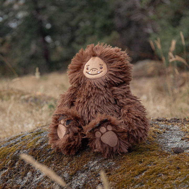 Sully the Sasquatch brown plush stuffed animal sitting on a mossy rock outdoors