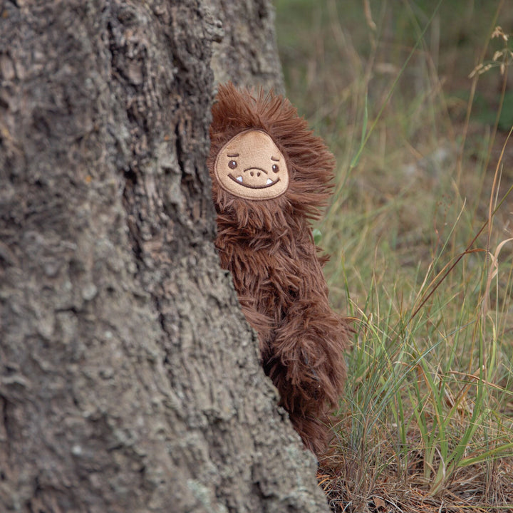 Sully the Sasquatch brown plush toy peeking from behind a tree outdoors