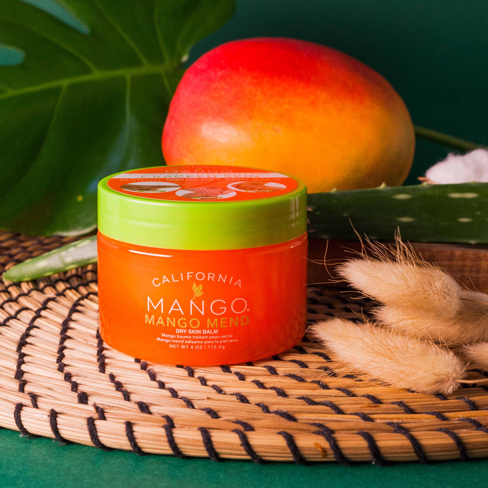 California Mango Mend Dry Skin Balm 4oz in orange jar with green lid surrounded by tropical props.