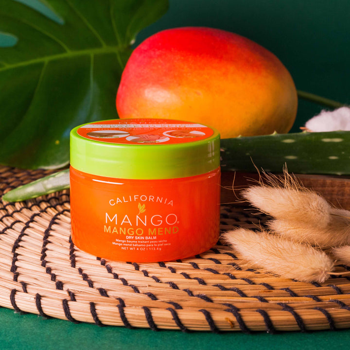 California Mango Mend Dry Skin Balm 4oz in orange jar with green lid surrounded by tropical props.