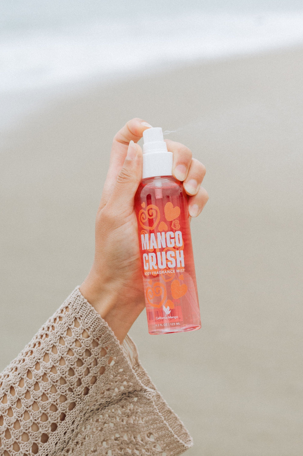 California Mango Crush Body Fragrance Mist 4oz spray bottle held up at a beach