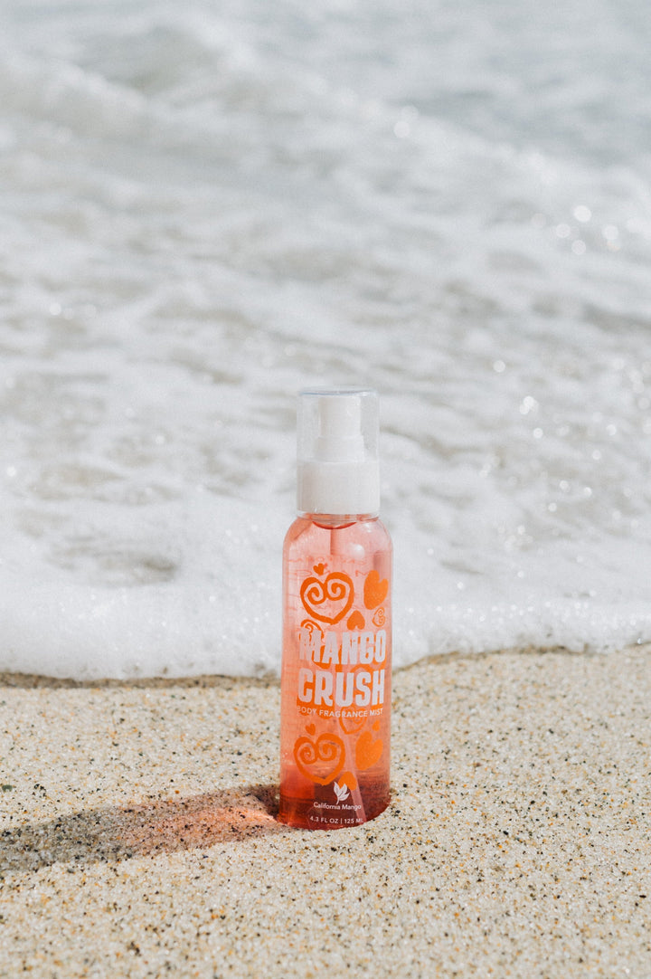 California Mango Crush Body Fragrance Mist 4oz spray bottle on sandy beach with ocean waves
