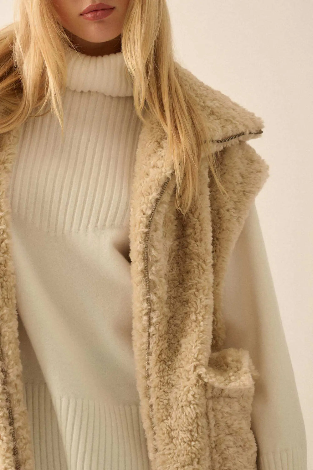 Woman wearing cream colored faux fur zip-up vest over white ribbed turtleneck sweater