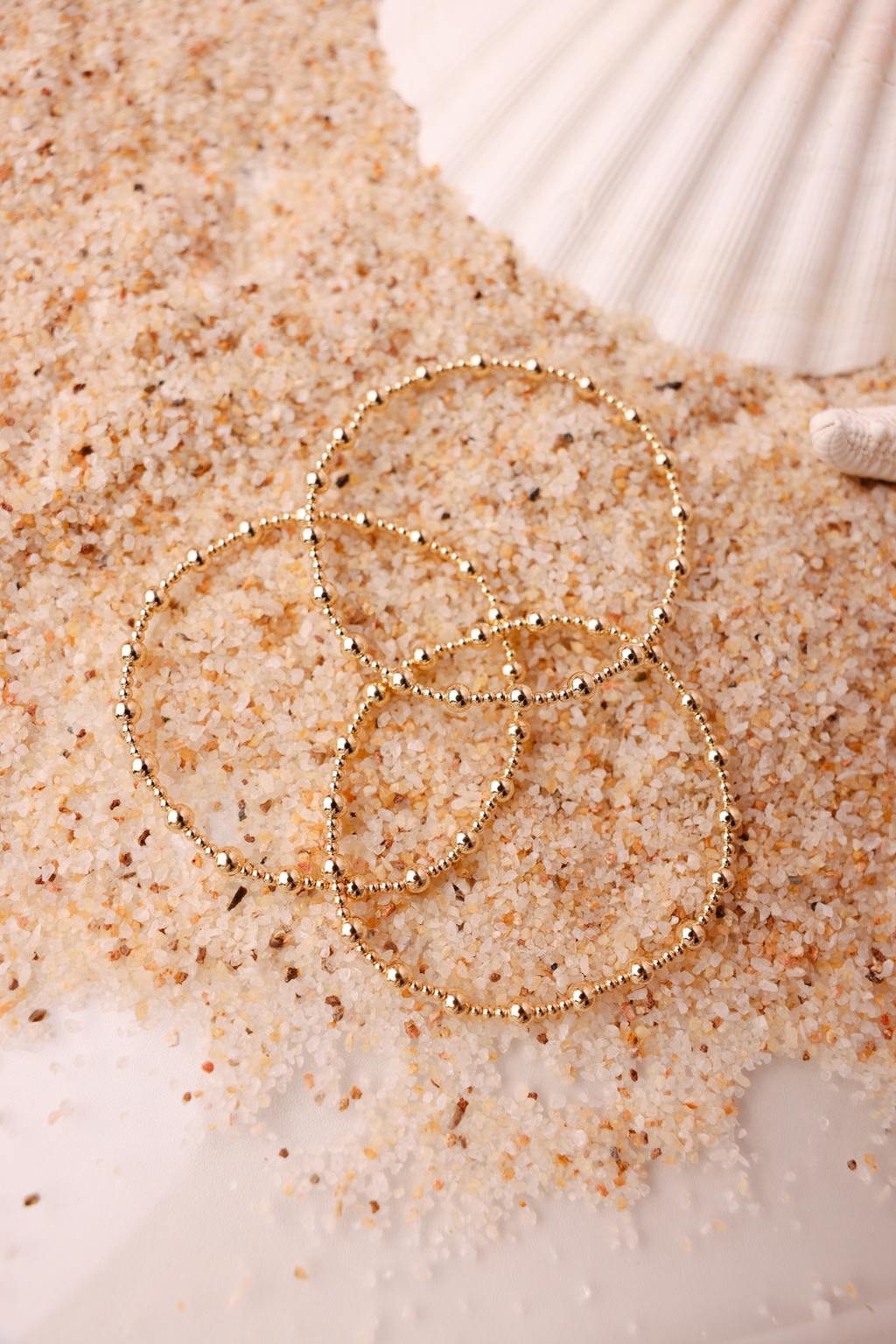 Three gold beaded stretch bracelets stacked on sandy beach surface with seashell backdrop