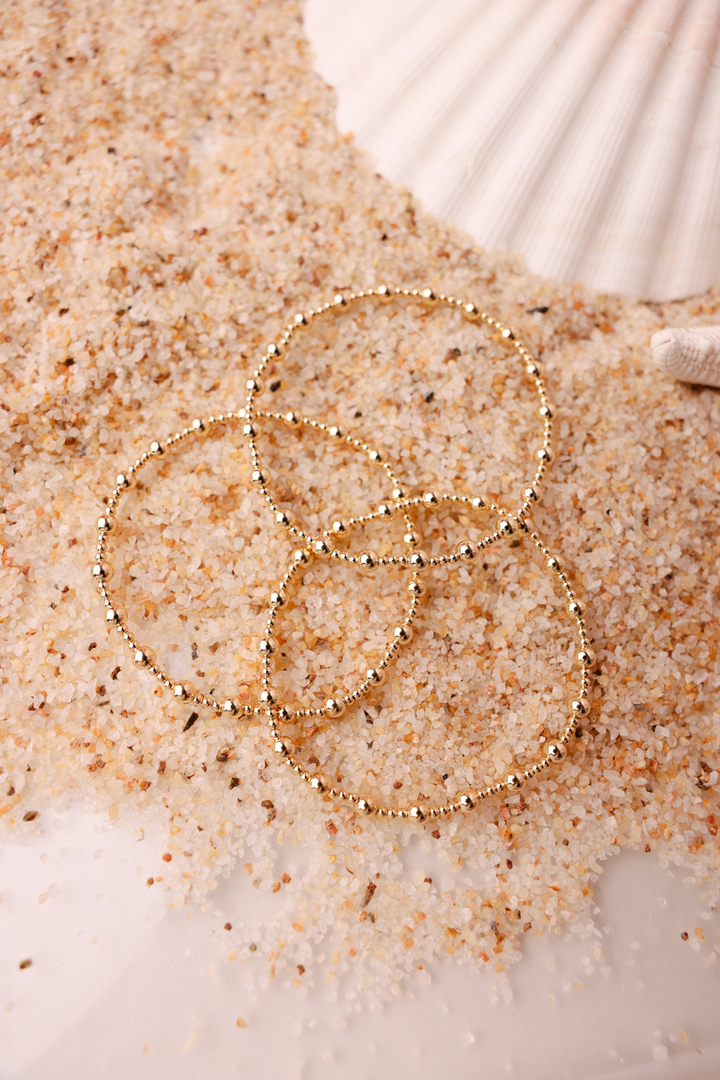 Three gold beaded stretch bracelets stacked on sandy beach surface with seashell backdrop