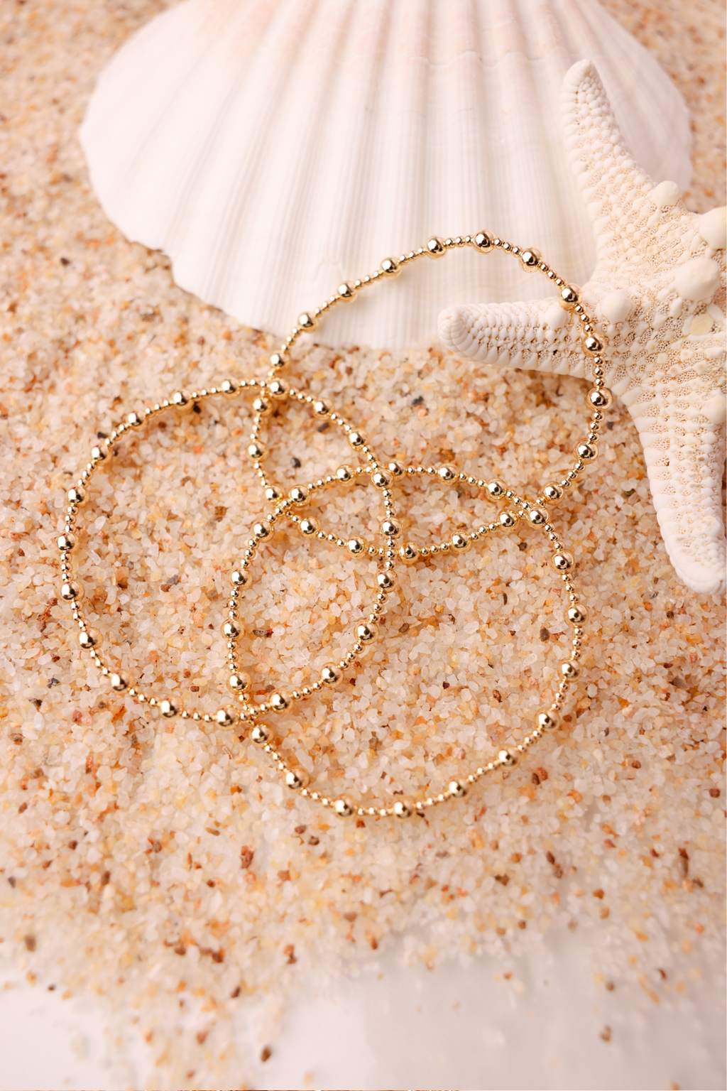 Beaded Blondes Palmer Bracelet in gold, delicate beaded chain coiled on sandy beach with shells.