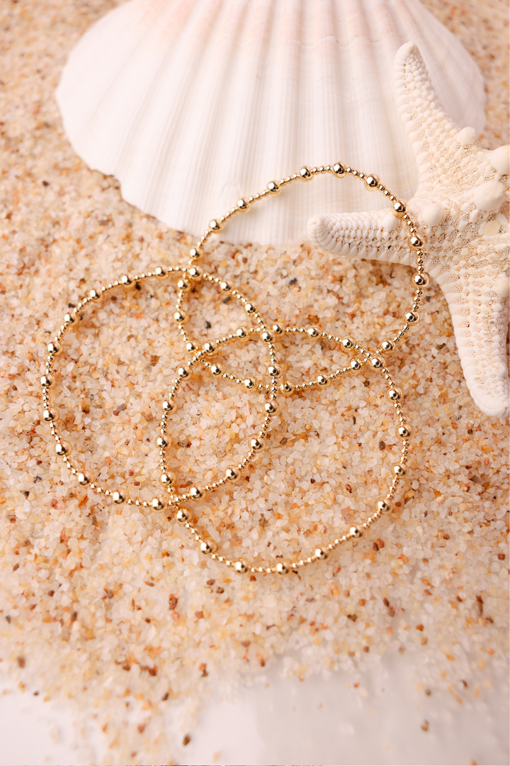 Beaded Blondes Palmer Bracelet in gold, delicate beaded chain coiled on sandy beach with shells.