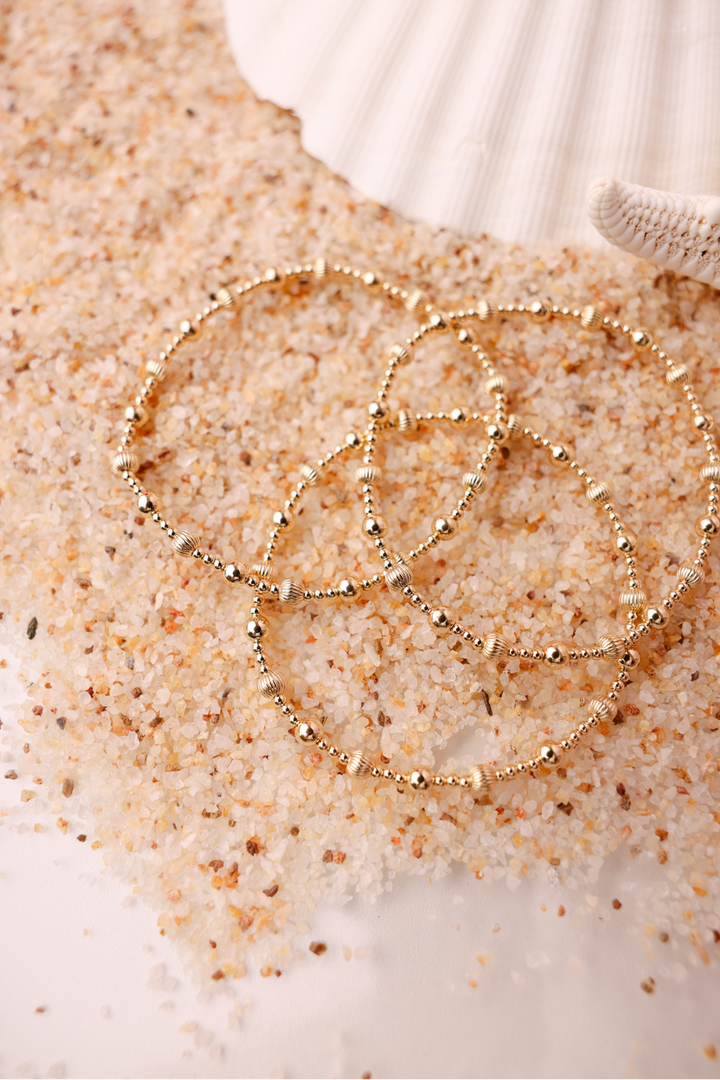 Three gold beaded bracelets with fluted accents displayed on sandy beach surface near seashell.