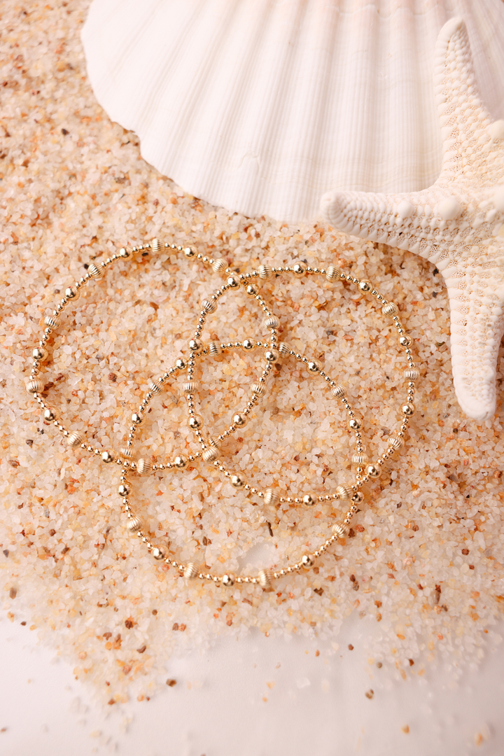 Gold beaded Palmer Fluted Bracelets displayed on sand with seashell and starfish backdrop.