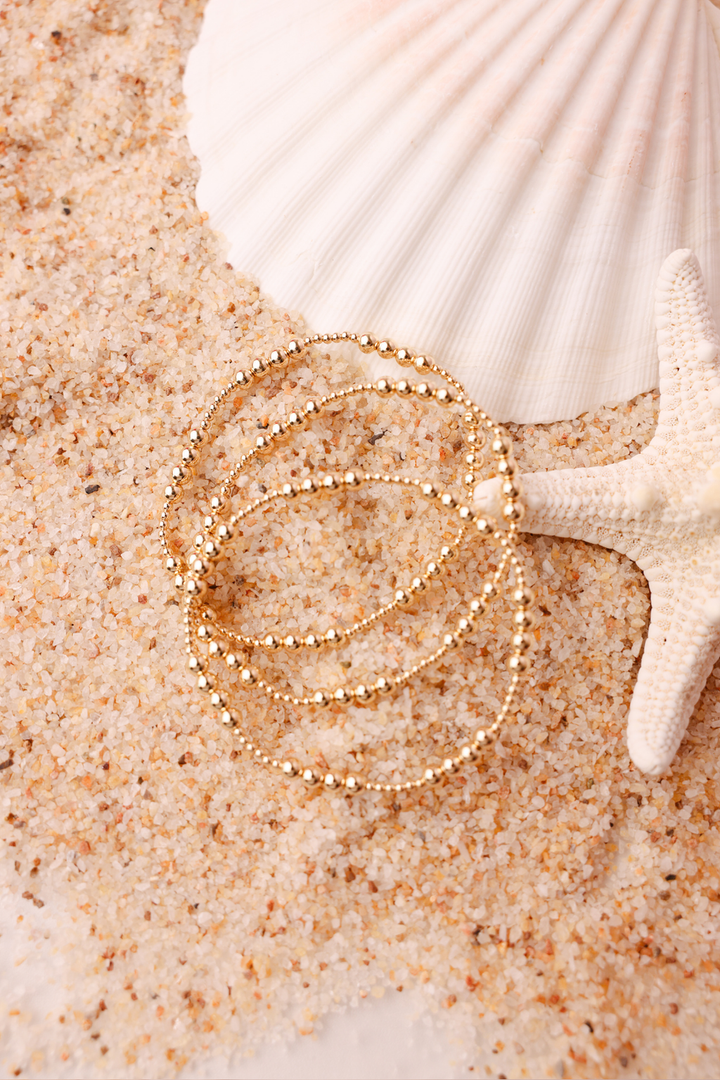 Beaded Blondes Scottie Bracelet in gold, stacked set displayed on sandy beach with seashells.