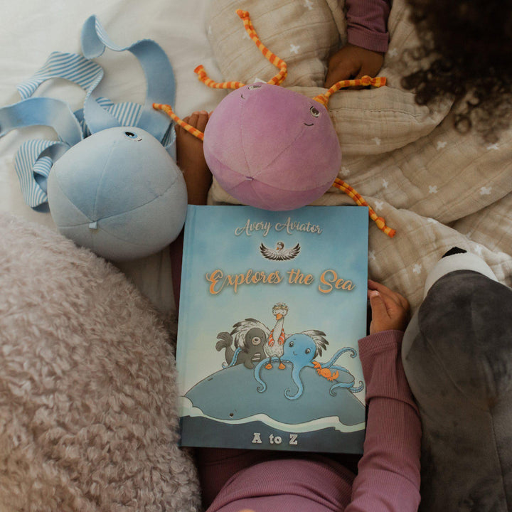 Child holding Avery Aviator Explores the Sea A to Z children's book with plush toys nearby