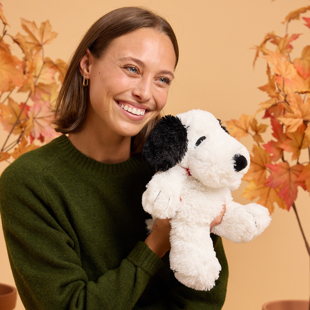 Woman holding white and black Snoopy Warmies plush toy with autumn leaves background
