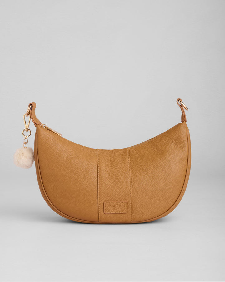 Tan leather crescent-shaped hobo bag with pom pom charm on neutral background