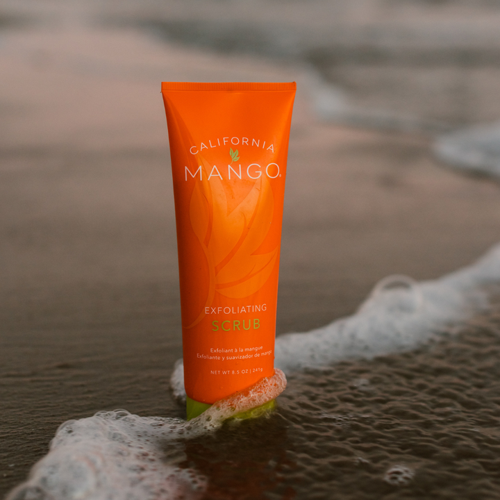 California Mango Exfoliating Scrub 8.5oz orange tube standing on a beach shoreline.