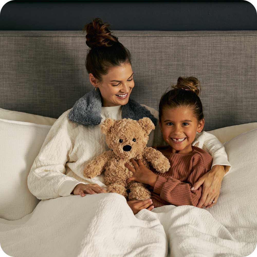 Mother and daughter cuddling with brown curly Warmies teddy bear on bed