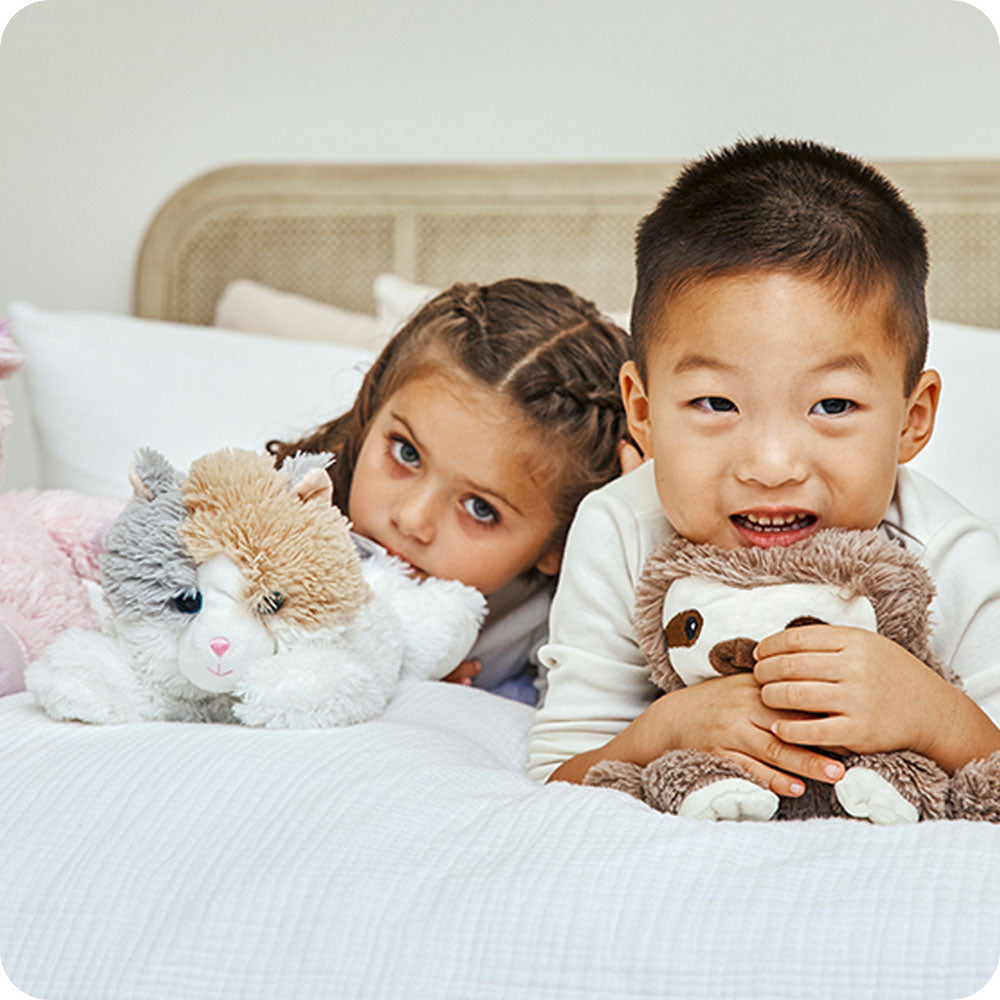 Two children in bed holding Warmies stuffed animals including calico cat plushie