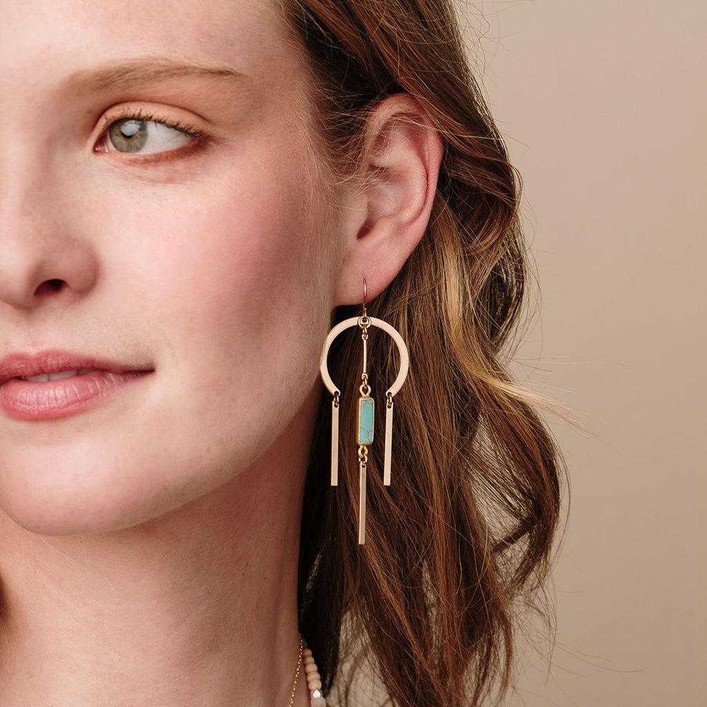 Woman wearing gold circular earrings with turquoise stone and dangling bar elements